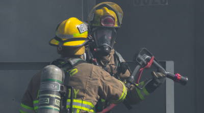 Two Unified Fire Authority firefighters communicate during a training scenario near a smoking house